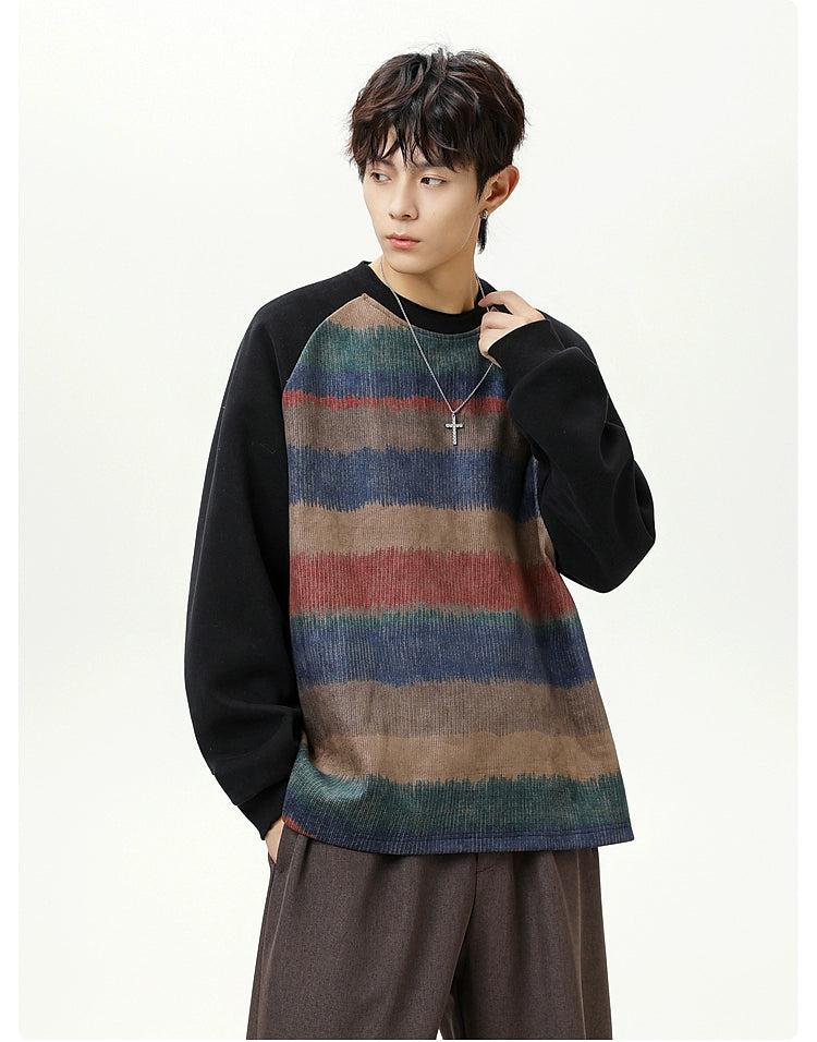 A young man wearing a striped sweater and a necklace stands against a white background.