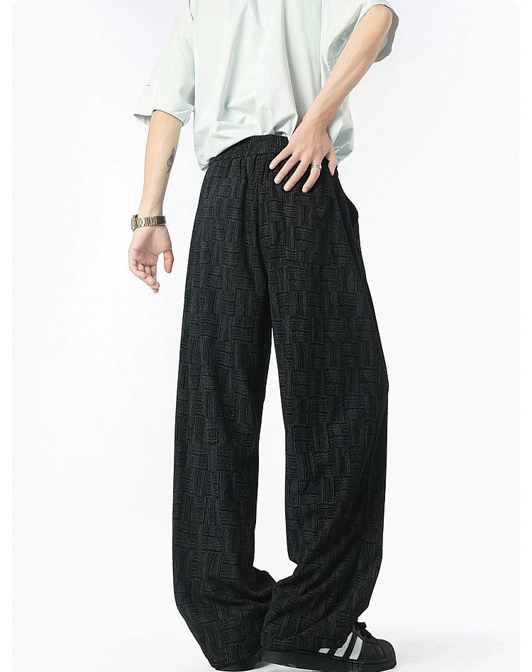 A person wearing a white shirt and black patterned pants stands with their hands in their pockets.