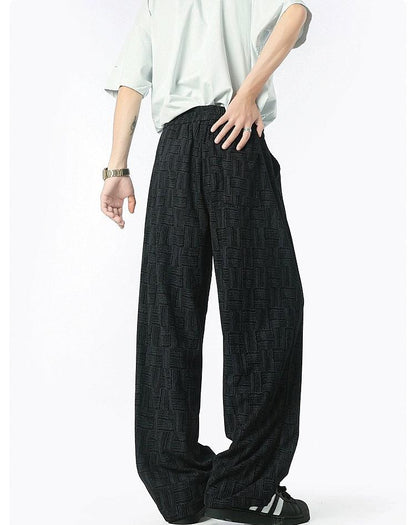 A person wearing a white shirt and black patterned pants stands with their hands in their pockets.