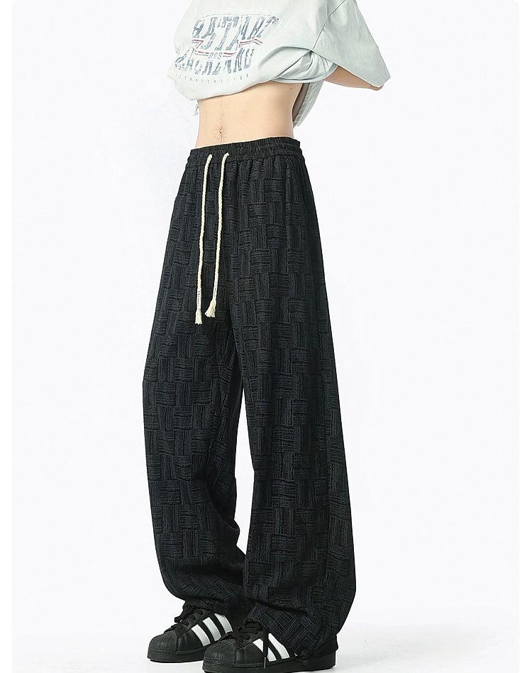 A person wearing a white shirt with a graphic design and black patterned pants stands against a white background.